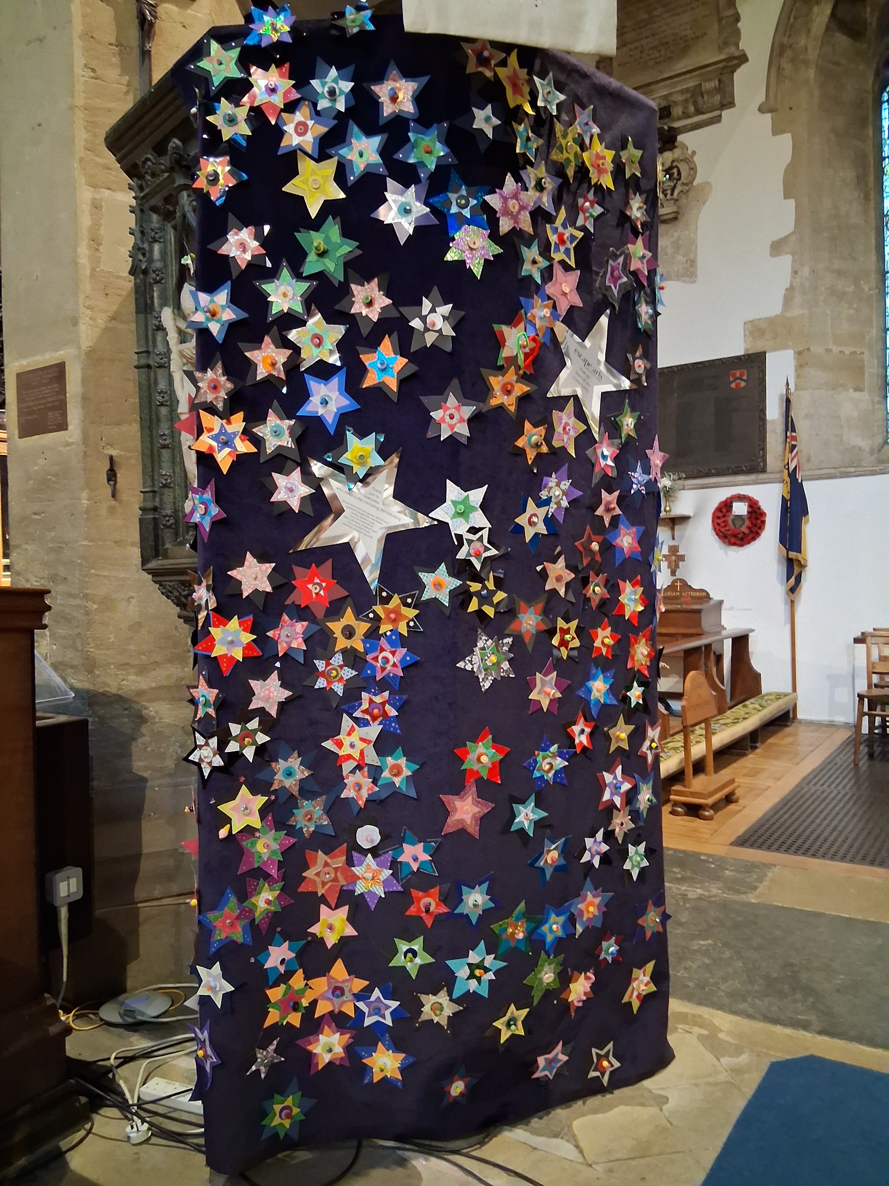 A tall black fabric panel covered with colourful, decorated paper stars and a few large silver stars stands inside a church, near wooden pews and stone walls.