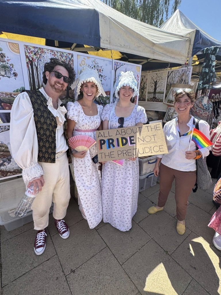 Four people stand at an outdoor event; three wear Regency-era costumes, one holds a rainbow flag. Another holds a sign reading All about the PRIDE, not the PREJUDICE. Art prints are displayed behind them.