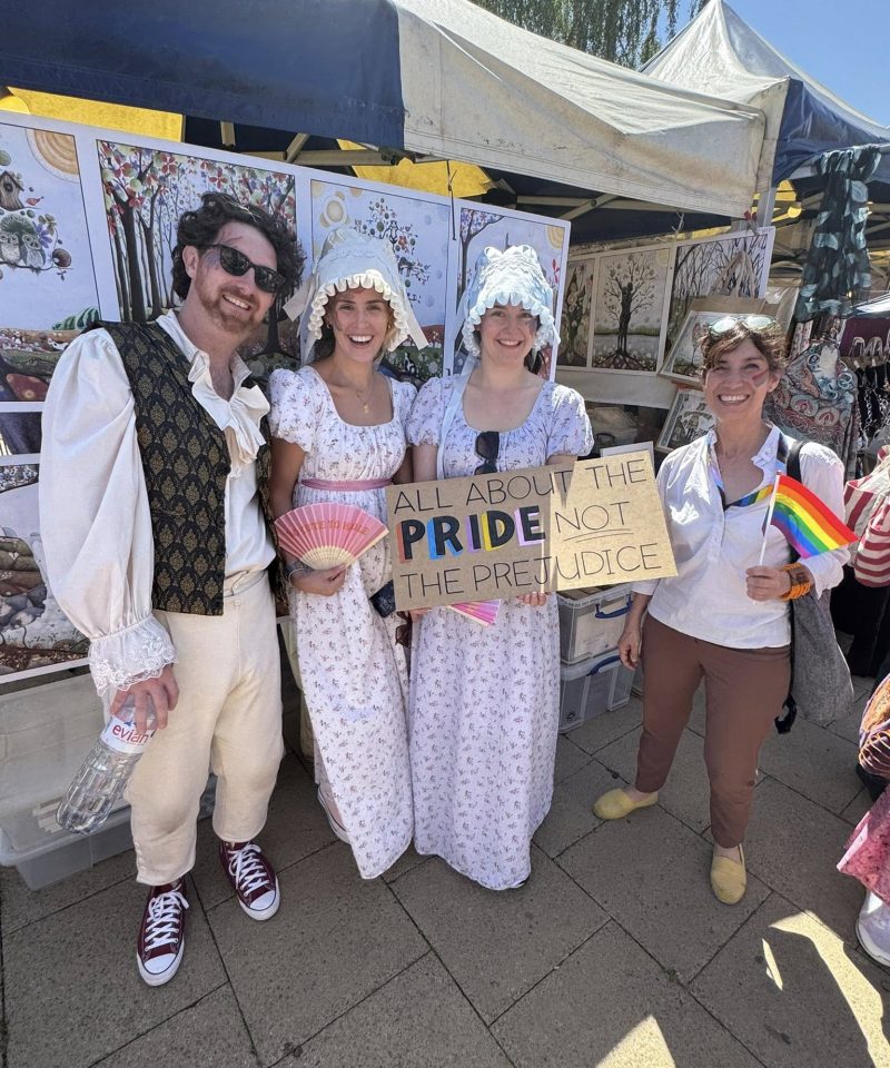 Four people stand at an outdoor event; three wear Regency-era costumes, one holds a rainbow flag. Another holds a sign reading All about the PRIDE, not the PREJUDICE. Art prints are displayed behind them.