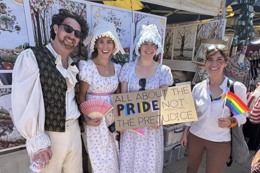Four people stand at an outdoor event; three wear Regency-era costumes, one holds a rainbow flag. Another holds a sign reading All about the PRIDE, not the PREJUDICE. Art prints are displayed behind them.