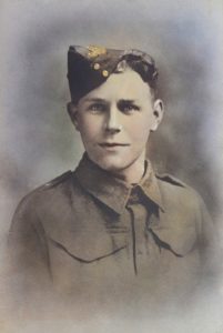 A young soldier in an old-fashioned military uniform and a side cap, looking directly at the camera. The image is colourised and has a soft, faded background.
