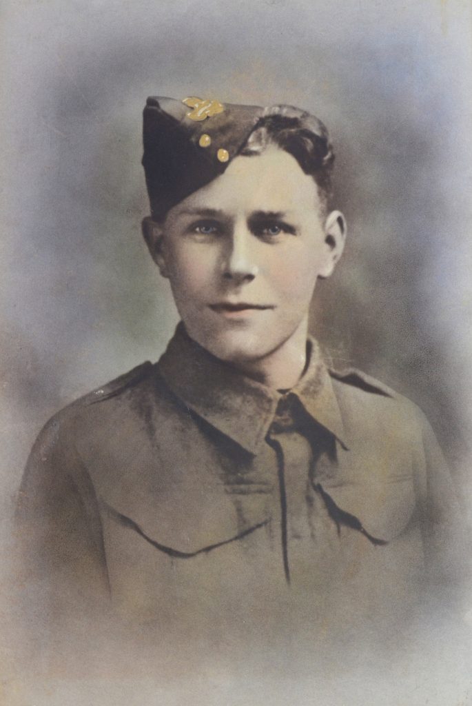 A young soldier in an old-fashioned military uniform and a side cap, looking directly at the camera. The image is colourised and has a soft, faded background.