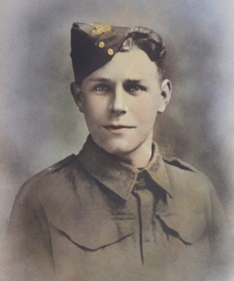 A young soldier in an old-fashioned military uniform and a side cap, looking directly at the camera. The image is colourised and has a soft, faded background.
