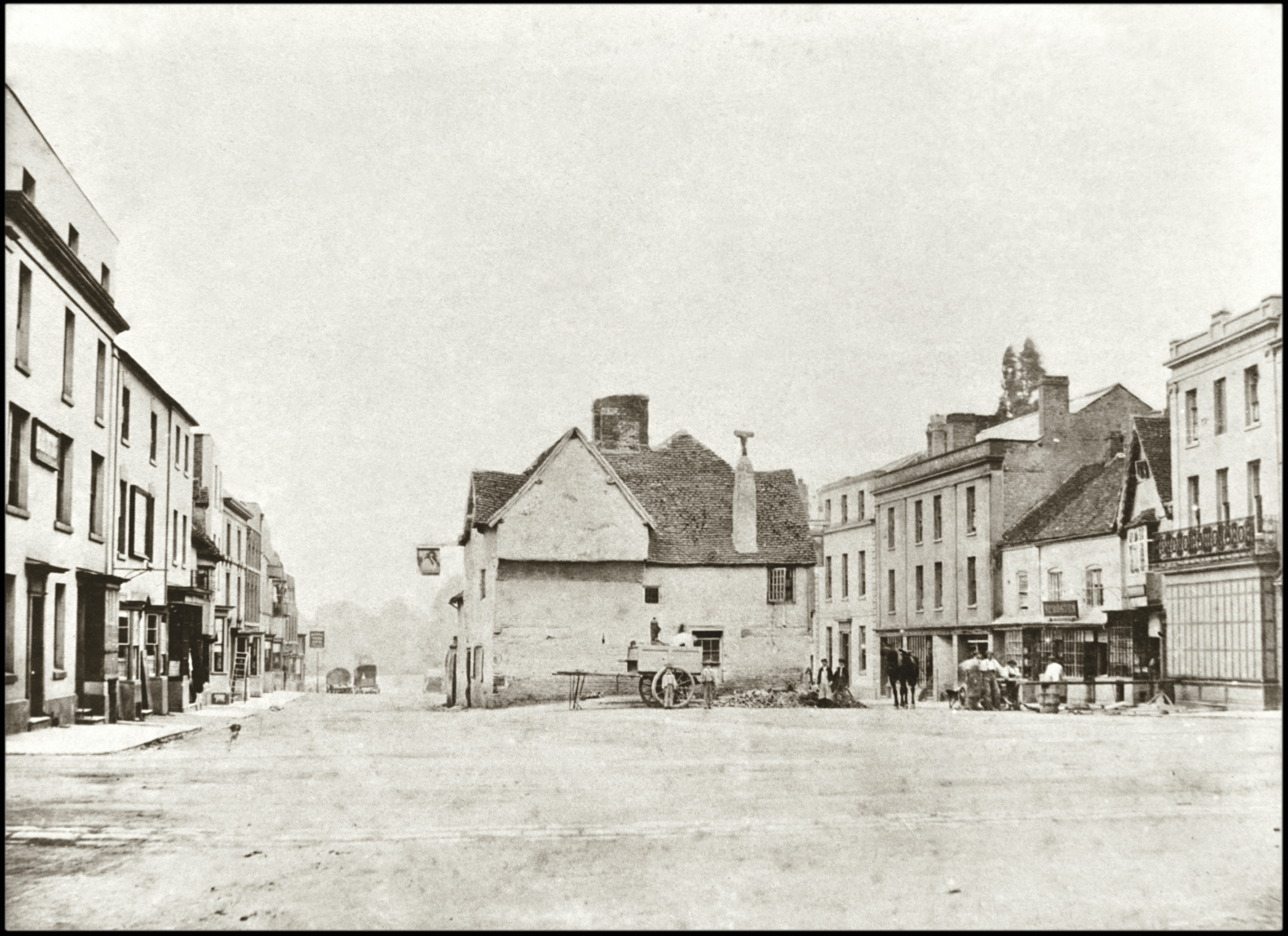 Old black-and-white photo of a quiet town square with a cart in front of a small central building, surrounded by larger buildings on both sides. There are a few people and horses in the background.