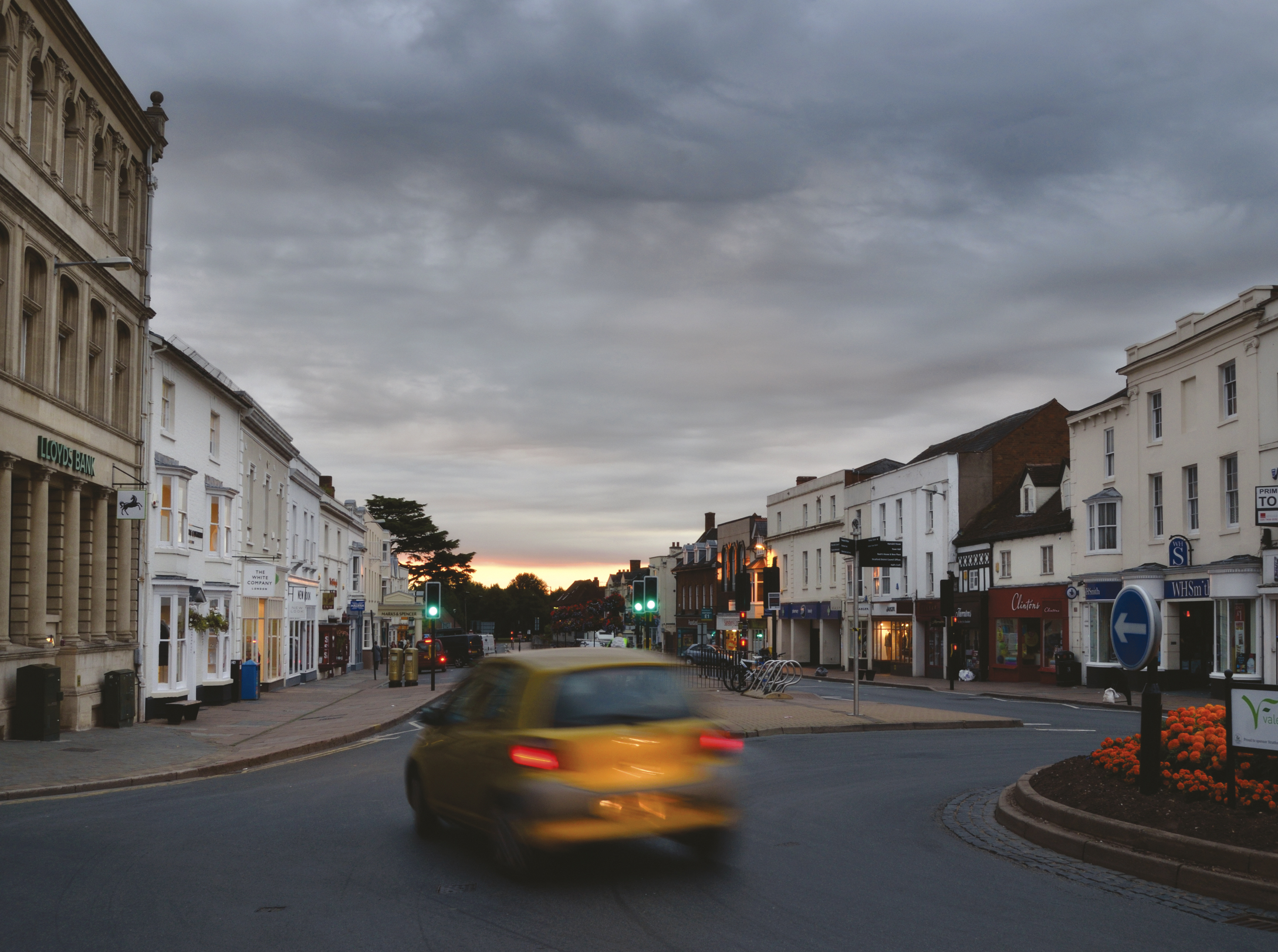 A yellow car drives through a roundabout in a quiet town at dusk, with white shops and buildings lining both sides of the street under a cloudy sky.