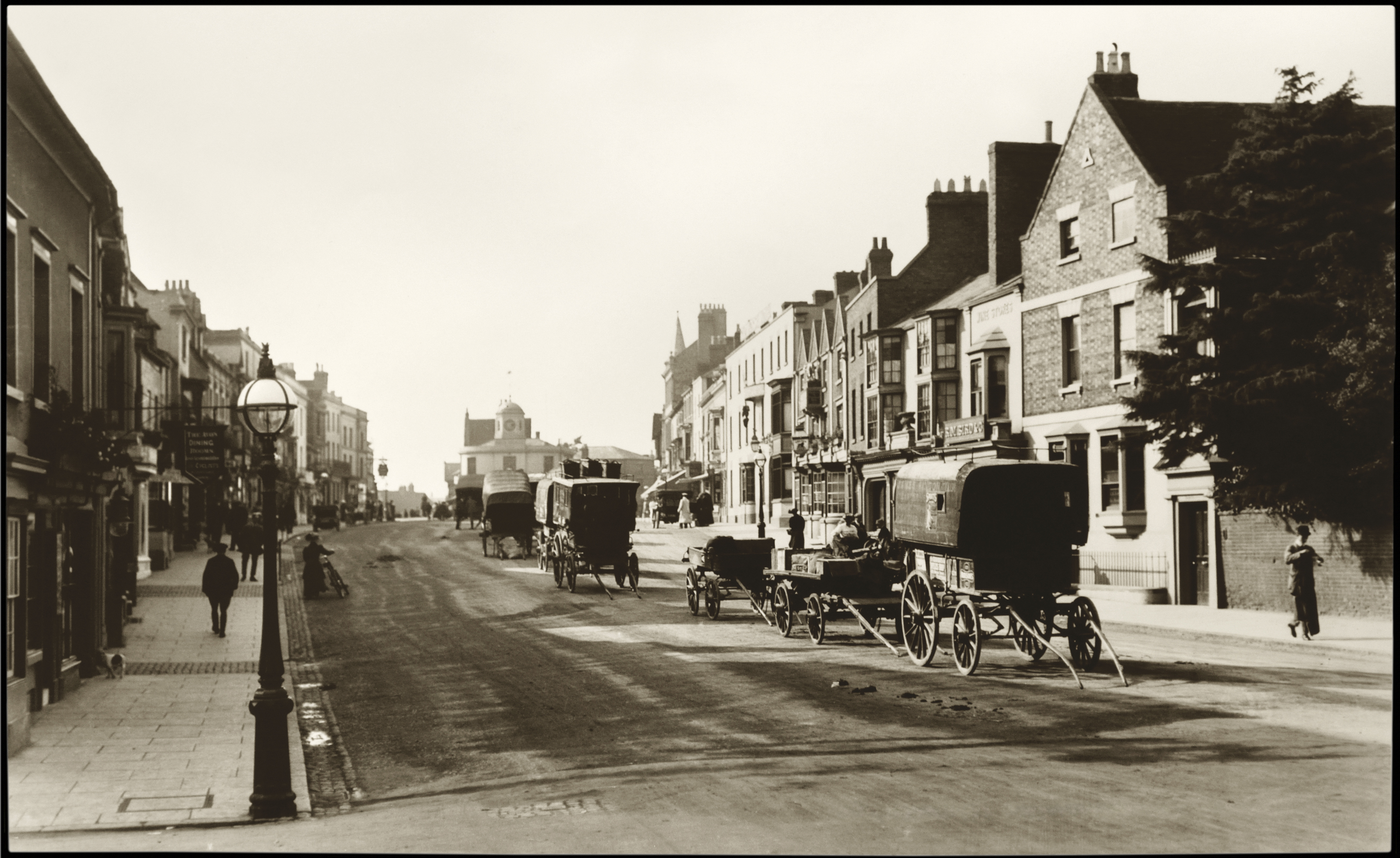 A sepia-toned photo of an old street lined with buildings, horse-drawn carriages parked along the road, and a few people walking on the pavements.