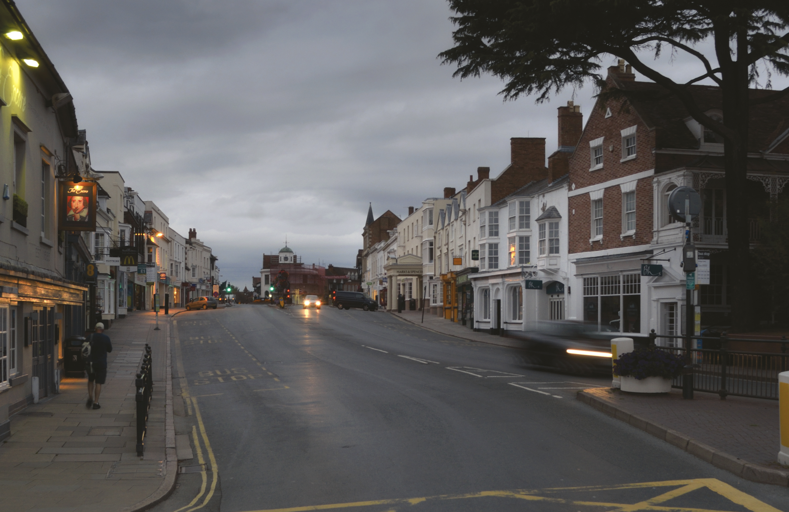 A quiet street lined with historic buildings under a cloudy evening sky, with a few pedestrians and blurred cars passing by.