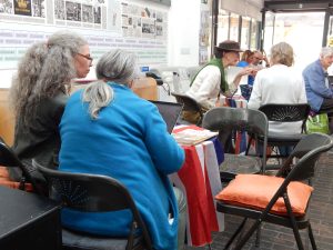 Several older adults sit around tables covered with a red, white, and blue cloth, talking and working on laptops and papers in a bright room with folding chairs and historical displays on the wall.