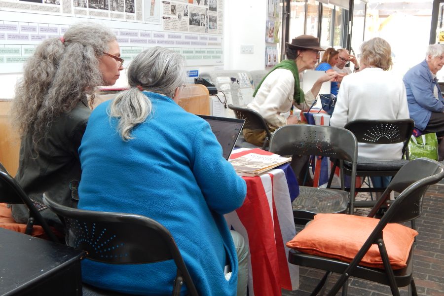 Several older adults sit around tables covered with a red, white, and blue cloth, talking and working on laptops and papers in a bright room with folding chairs and historical displays on the wall.