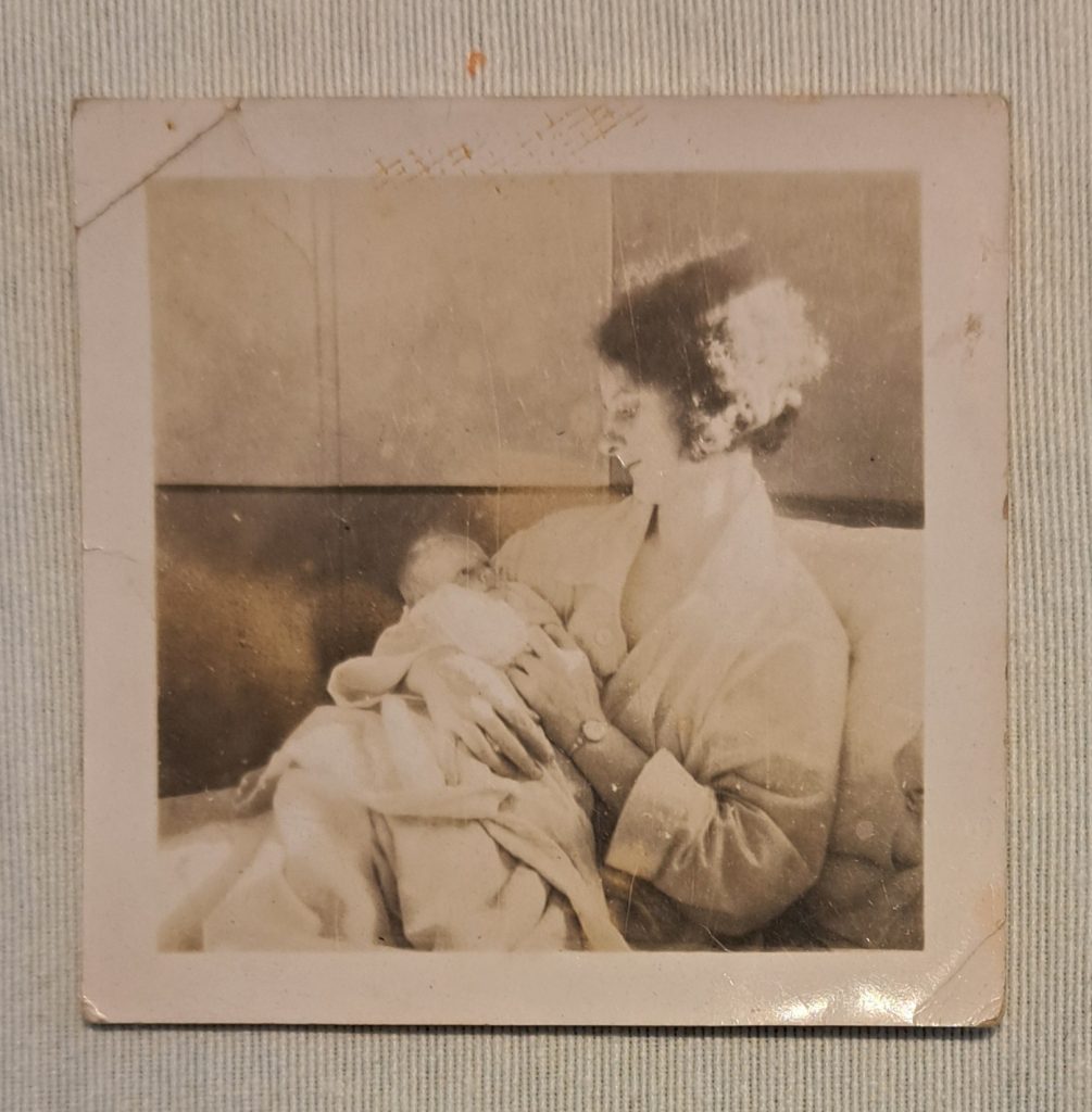 A vintage black and white photo shows a woman sitting and holding a newborn baby, both wrapped in blankets, with the woman gazing lovingly at the infant.