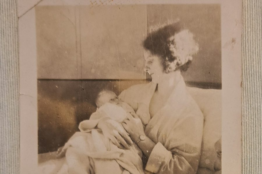 A vintage black and white photo shows a woman sitting and holding a newborn baby, both wrapped in blankets, with the woman gazing lovingly at the infant.