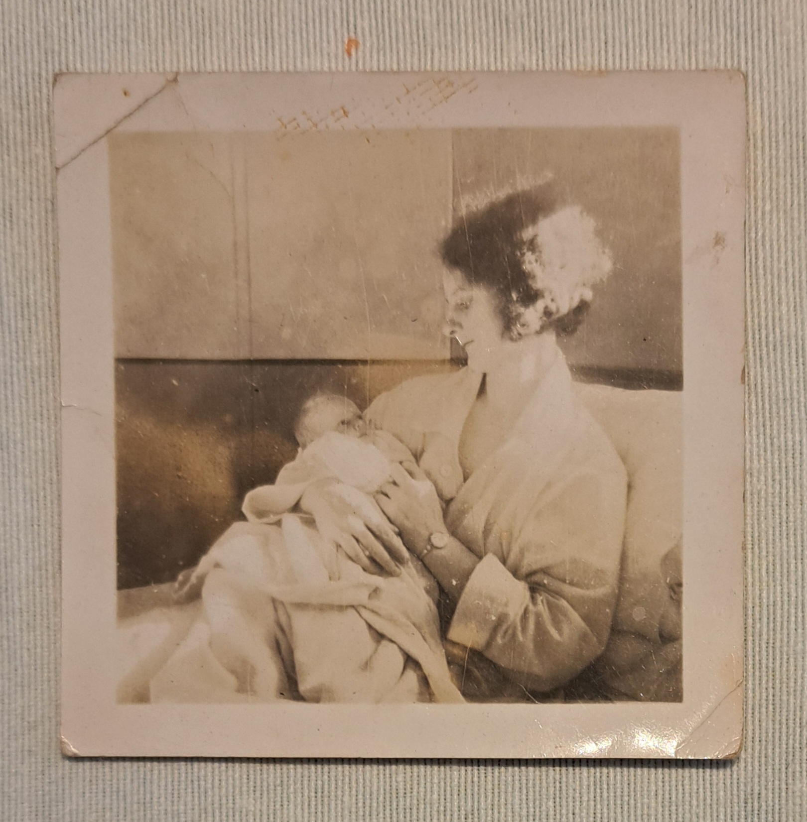 A vintage black and white photo shows a woman sitting and holding a newborn baby, both wrapped in blankets, with the woman gazing lovingly at the infant.