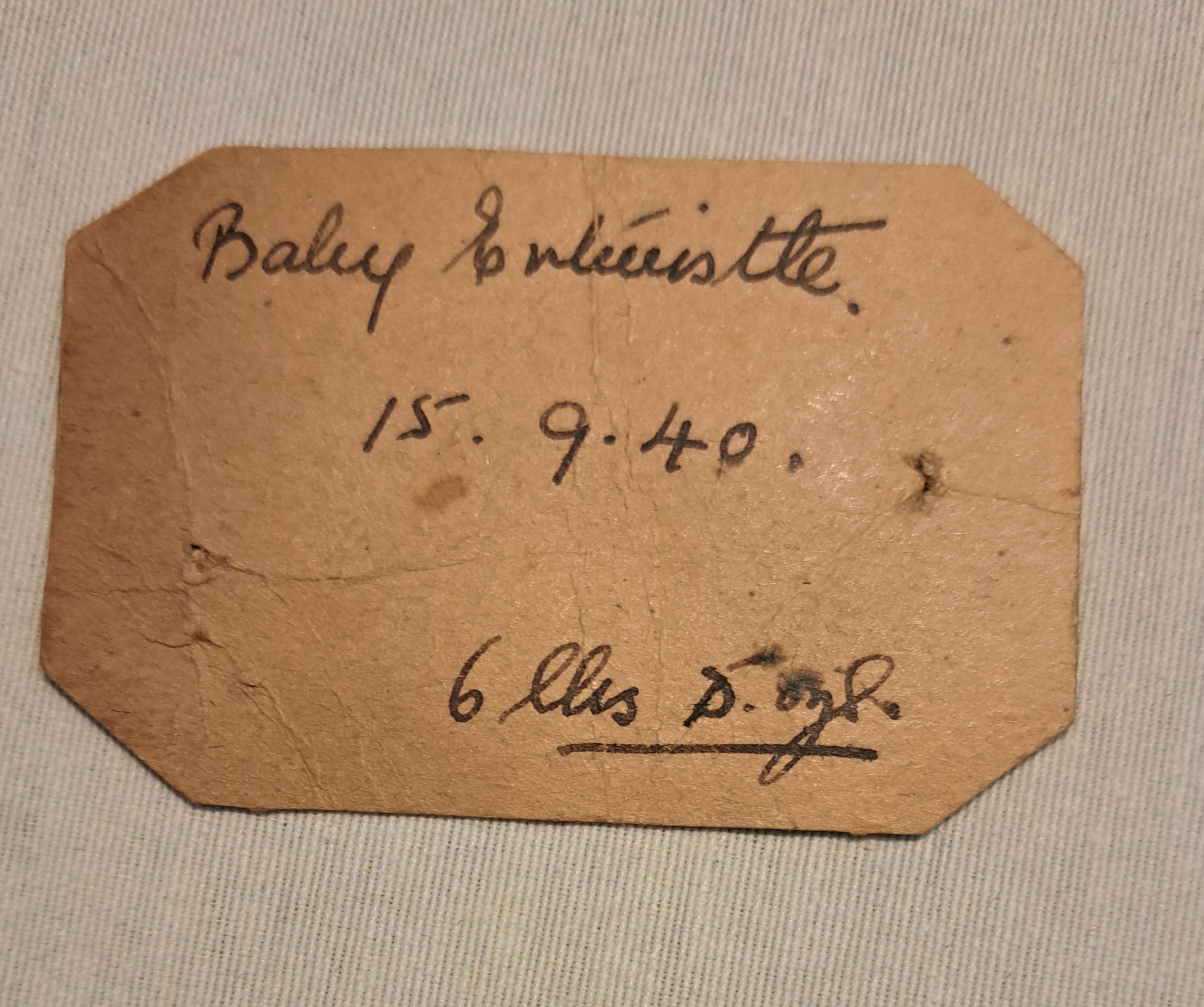 A brown, worn paper label with handwritten text: Baby Spinister. 15.9.40. 6 lbs 8 oz. on a light striped fabric background.