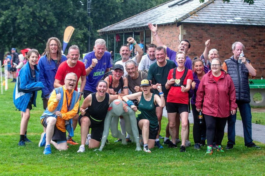 A group of people, some in sportswear, pose and smile together outdoors on a rainy day, with a brick building and wet grass in the background. Some hold medals and show celebratory gestures.
