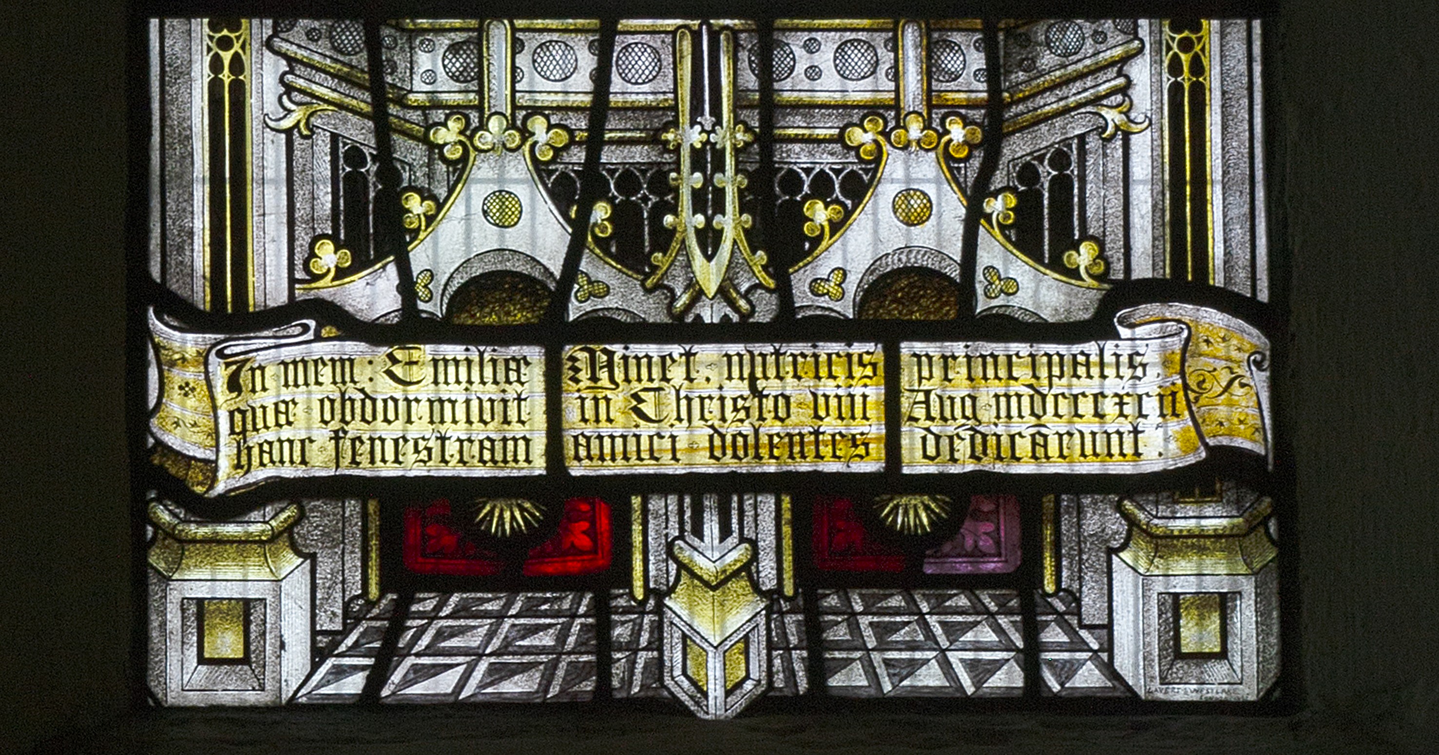 Stained glass window with ornate patterns, arches, and Latin text on a yellow scroll in the foreground, set against a geometric floor and architectural background.