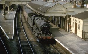 A vintage steam locomotive numbered 5337 is stopped at a railway station platform, with cream and brown station buildings and tracks curving away. Few people are visible on the platform.