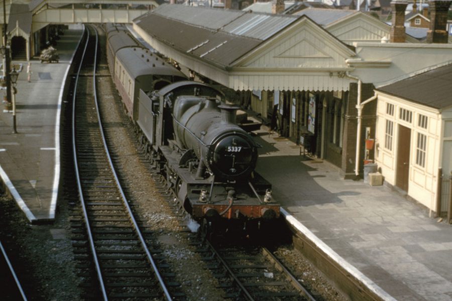 A vintage steam locomotive numbered 5337 is stopped at a railway station platform, with cream and brown station buildings and tracks curving away. Few people are visible on the platform.