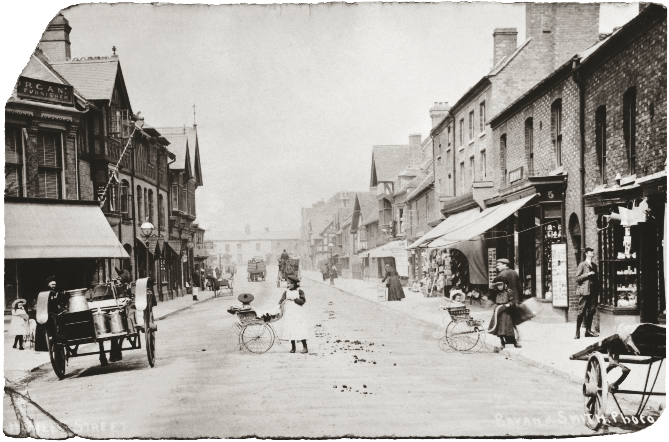 Black-and-white photo of a busy, old-fashioned street with children playing, women pushing prams, shops on both sides, and a horse-drawn cart in the background. The scene appears to be from the early 20th century.