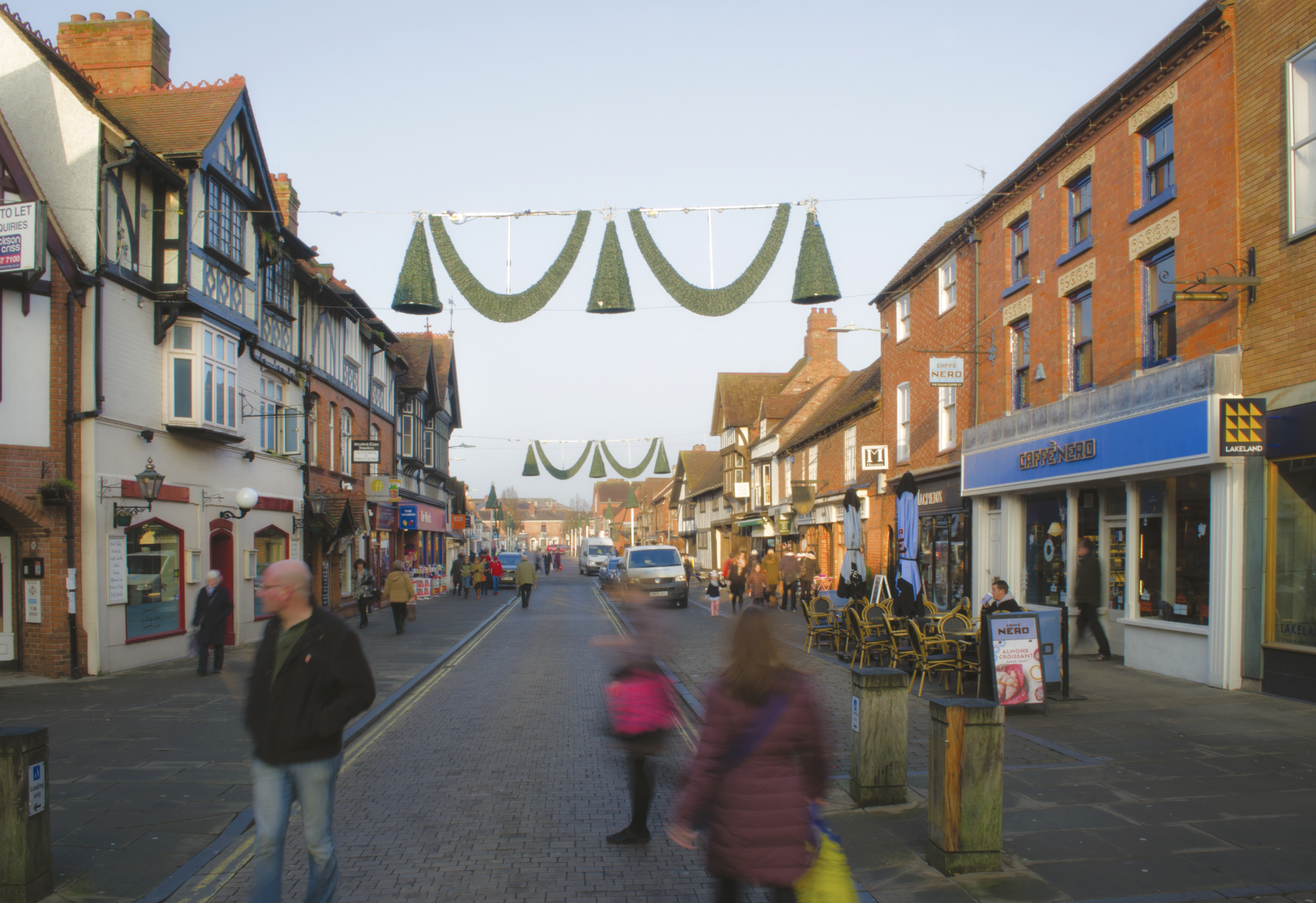 A busy street scene with people walking, historic buildings on both sides, outdoor seating, and green decorations hanging above the road on a clear day.