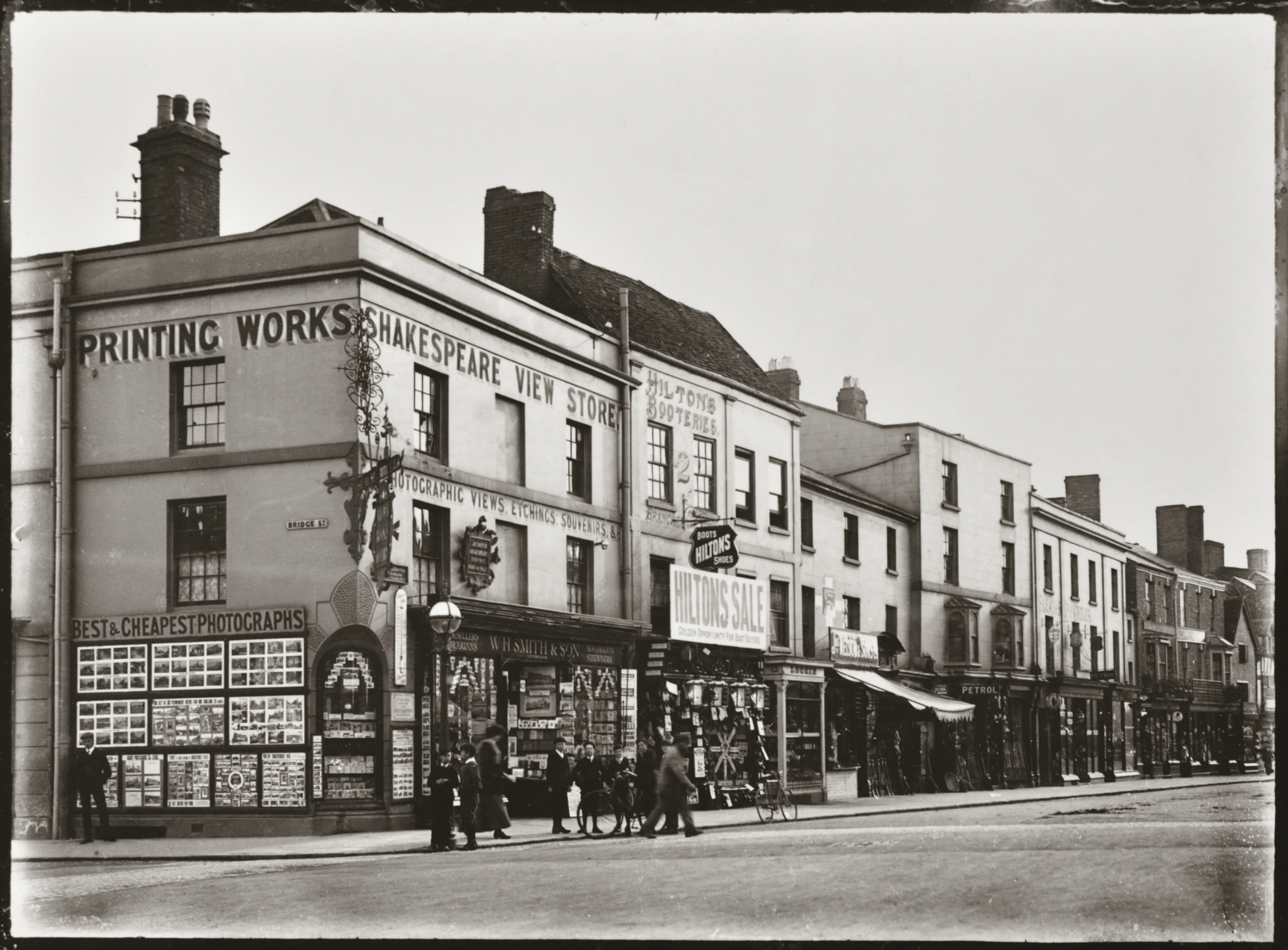 A black and white photo of a busy street with shops, including Printing Works and Shakespeare View Stores. People and bicycles are in front, and posters cover the walls. The scene appears to be from the early 1900s.