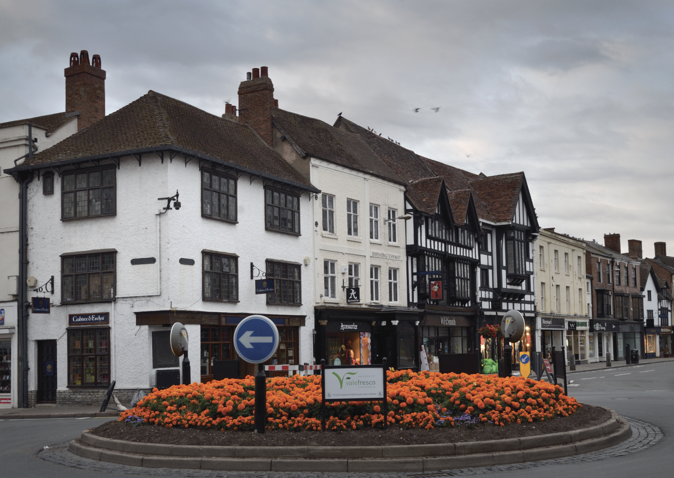 Historic buildings with timber frames line a street behind a roundabout filled with orange flowers; shops and signs are visible under a cloudy sky.