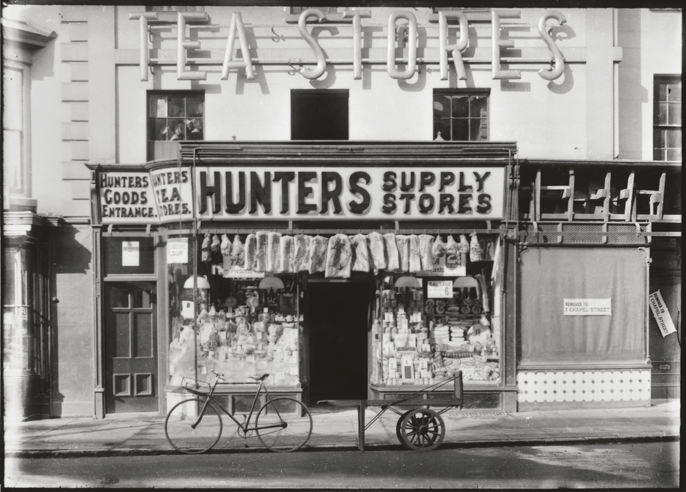 Black and white photo of Hunters Supply Stores, a shop with goods displayed in the window, a bicycle cart out front, and large signs above reading TEA STORES and HUNTERS SUPPLY STORES.