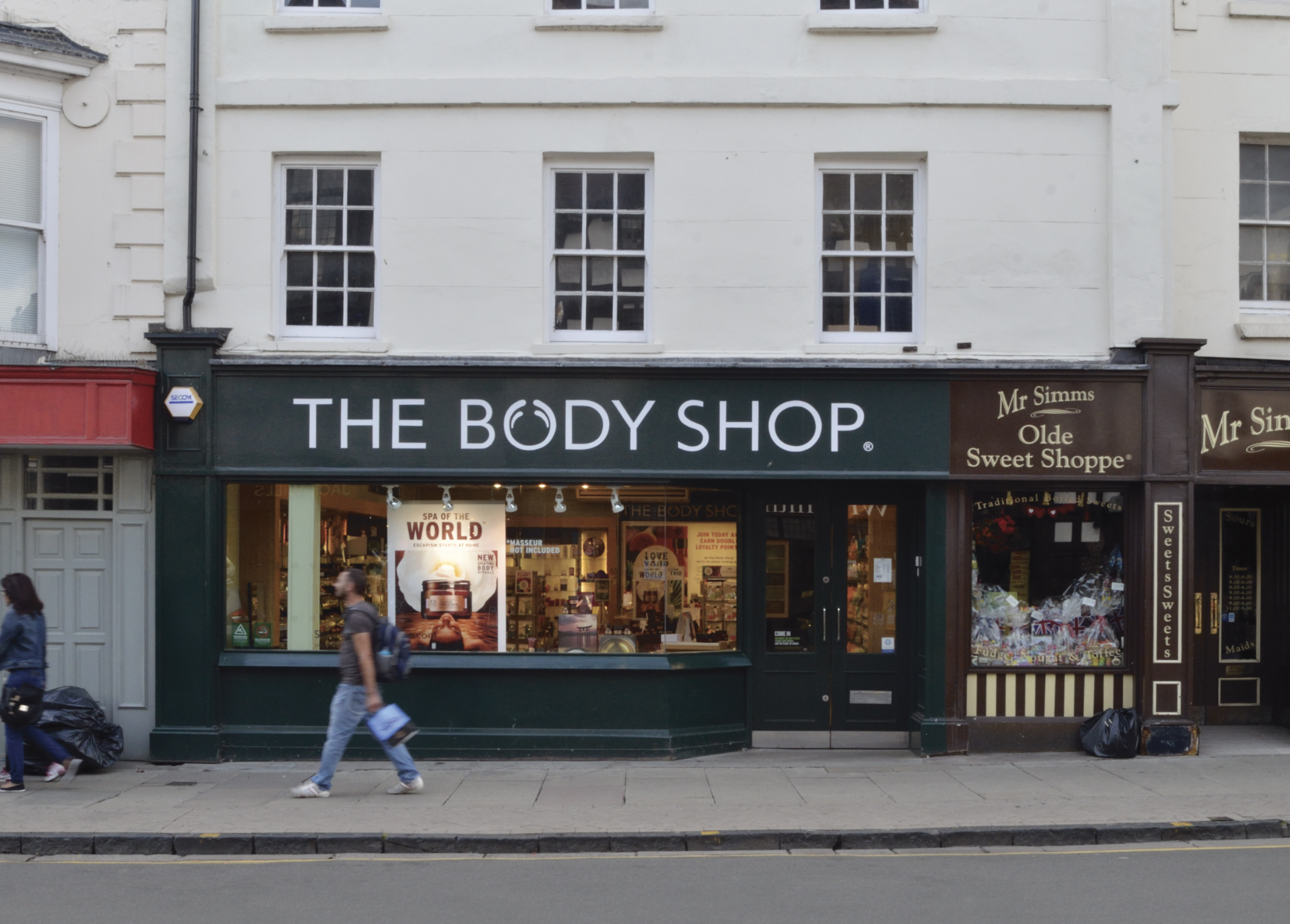 A person walks past The Body Shop shop, which has large glass windows displaying products. Adjacent shops include a sweet shop with jars and sweets visible inside. The street is quiet with minimal foot traffic.