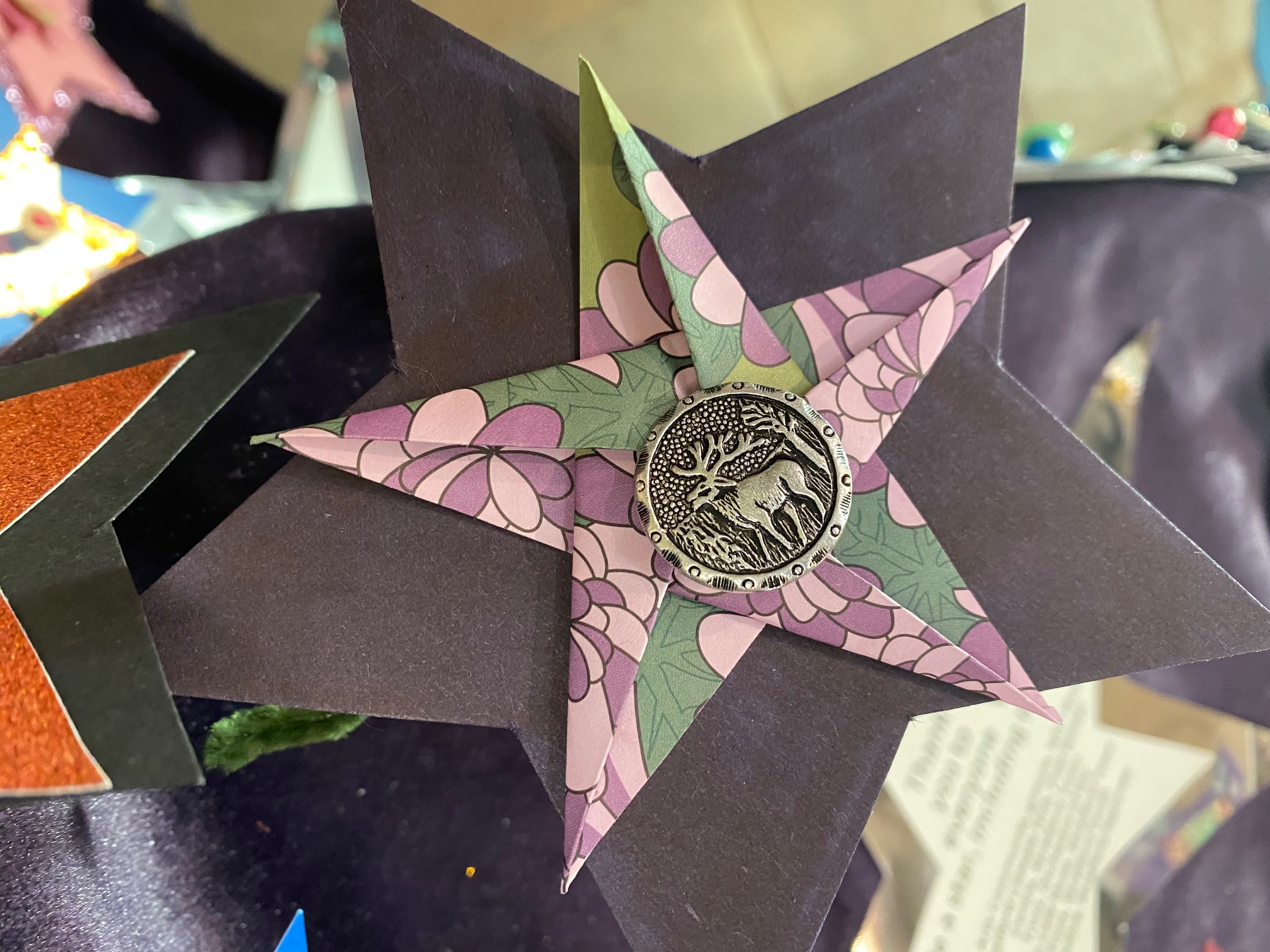 A black and purple origami star flower with a vintage coin featuring a face at its centre, set against a blurred background with papers and other crafts.