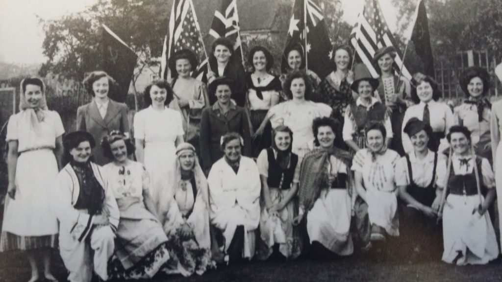A group of women in various traditional and historical costumes pose outdoors, some holding British, Australian, and American flags, in a black and white photograph.