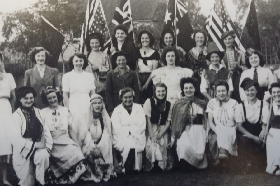 A group of women in various traditional and historical costumes pose outdoors, some holding British, Australian, and American flags, in a black and white photograph.