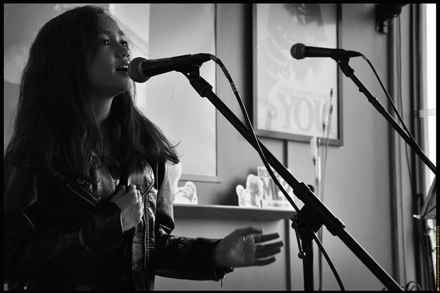 A young woman in a leather jacket sings into a microphone indoors, with another empty microphone nearby and posters on the wall in the background.