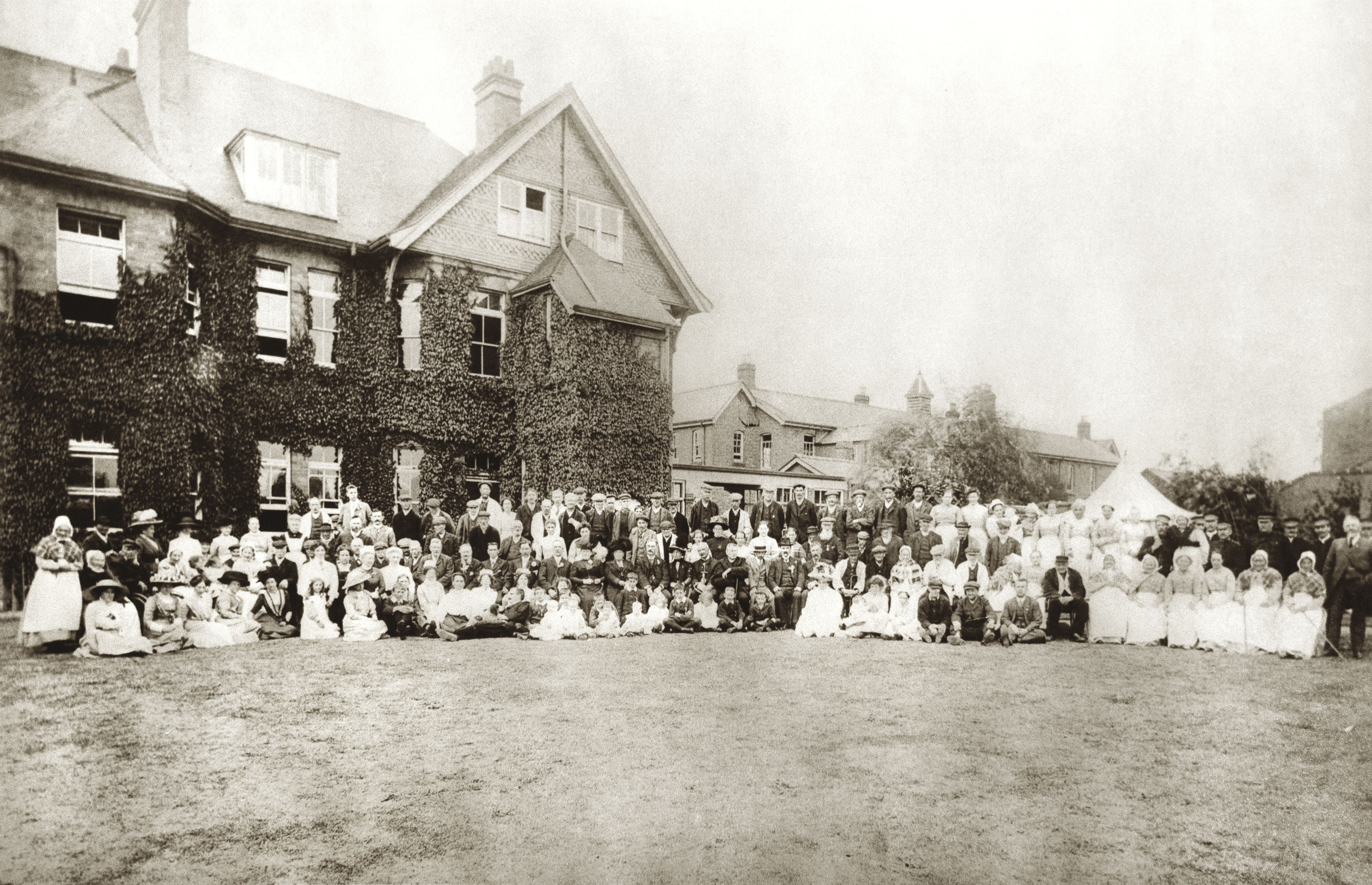A large group of men, women, and children pose in front of an ivy-covered brick building with multiple windows and chimneys, likely from the late 19th or early 20th century.