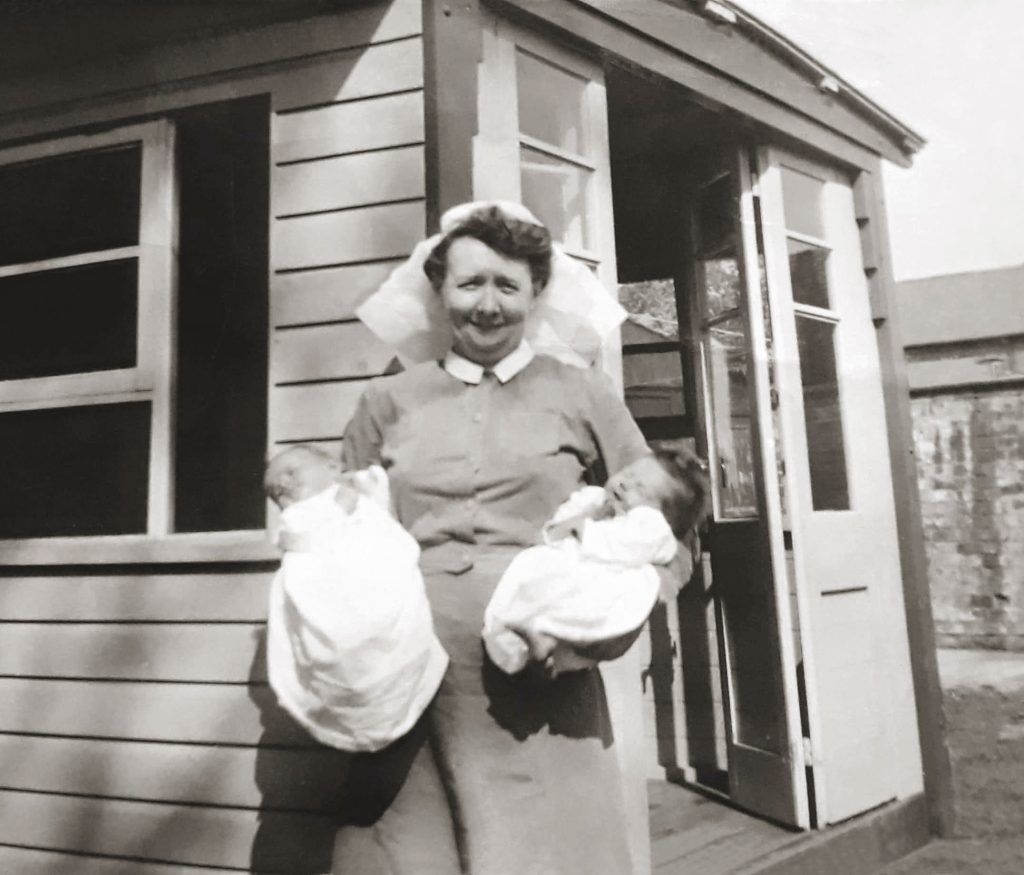 A nurse in a uniform and cap stands outside a wooden building, smiling and holding two swaddled newborn babies, one in each arm.