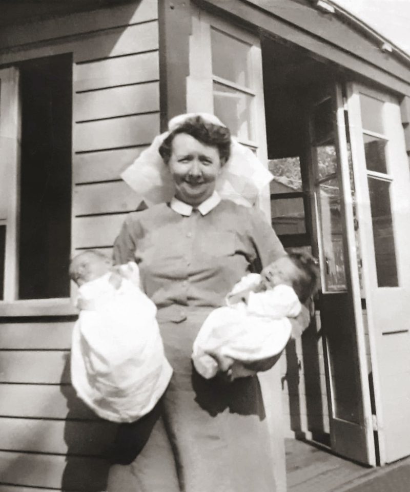 A nurse in a uniform and cap stands outside a wooden building, smiling and holding two swaddled newborn babies, one in each arm.