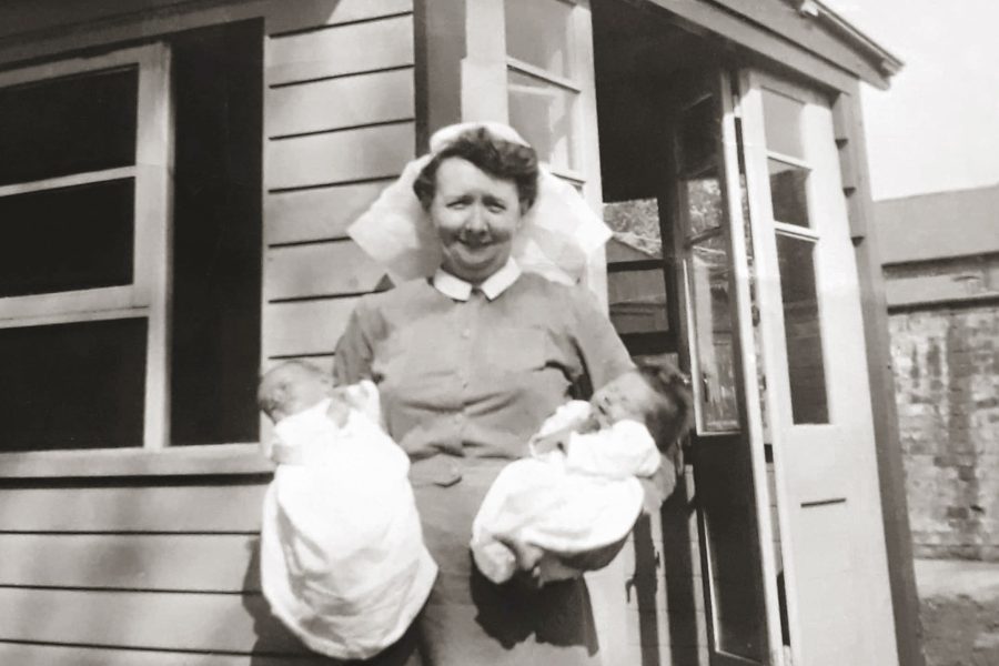 A nurse in a uniform and cap stands outside a wooden building, smiling and holding two swaddled newborn babies, one in each arm.