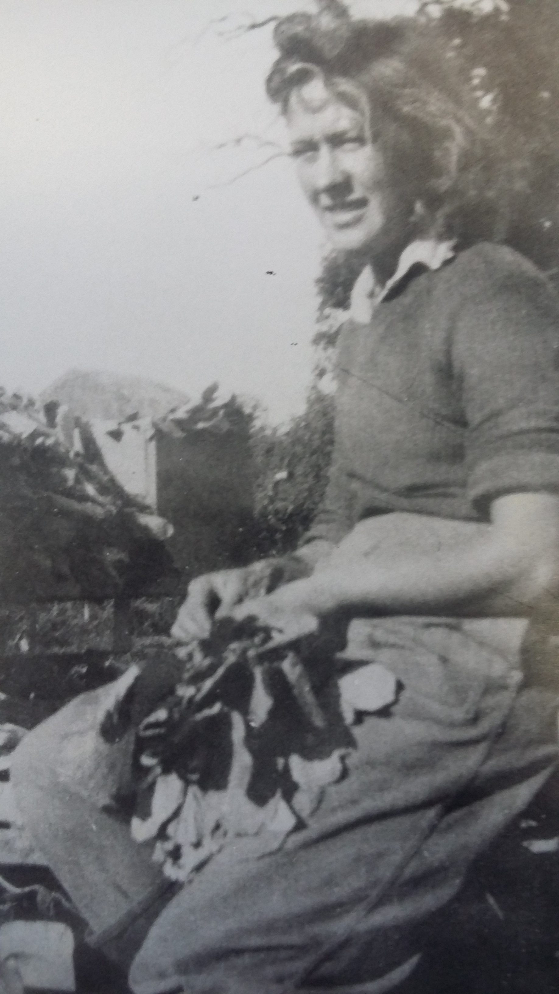 A young woman sits outdoors, holding leafy vegetables in her lap. She wears a jumper and trousers, with her hair styled in loose curls. The photo is black and white, and the background shows foliage.