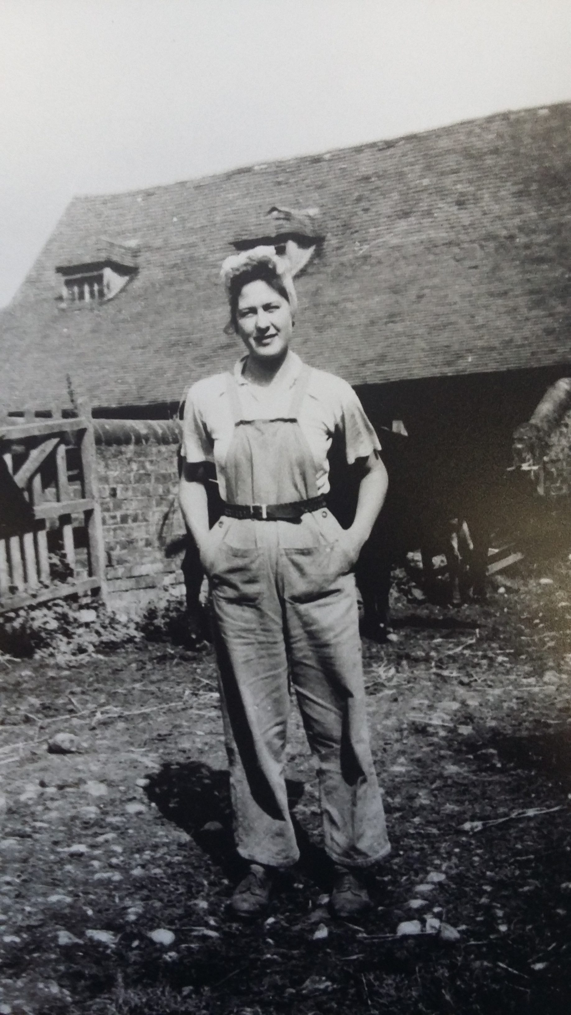 A person in overalls and a hat stands outside on bare earth, smiling, with hands behind their back. A wooden fence and rustic building are visible in the background.