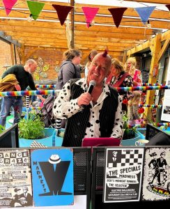 A man with a red mohican and polka-dot shirt sings into a microphone at an outdoor event, surrounded by people, colourful bunting, and posters for ska and punk bands.