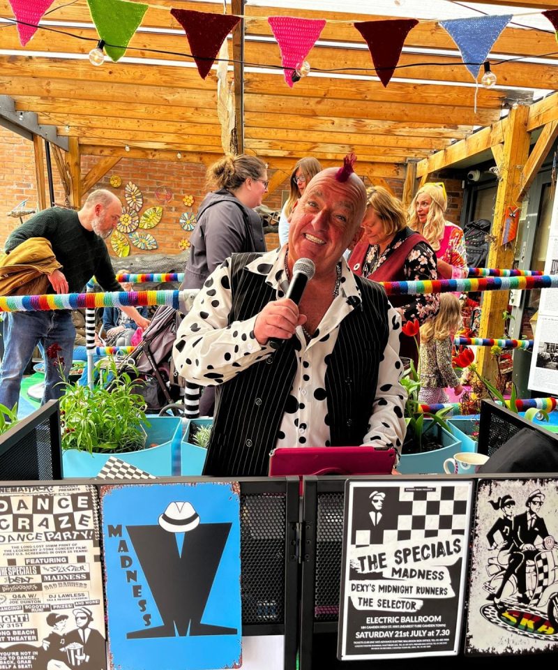 A man with a red mohican and polka-dot shirt sings into a microphone at an outdoor event, surrounded by people, colourful bunting, and posters for ska and punk bands.