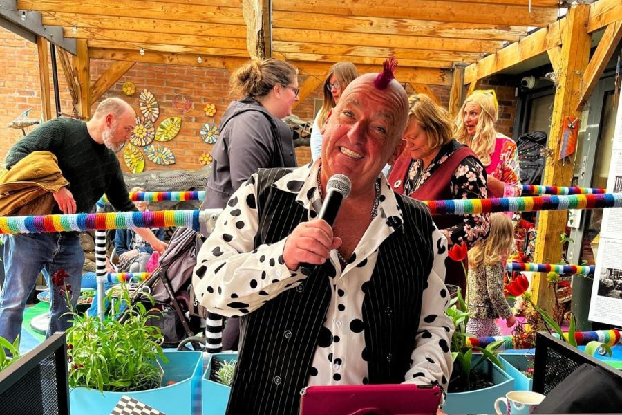 A man with a red mohican and polka-dot shirt sings into a microphone at an outdoor event, surrounded by people, colourful bunting, and posters for ska and punk bands.