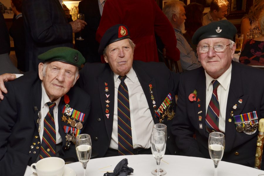 Three elderly men in military uniforms with medals and berets sit closely together at a table with champagne glasses, smiling at a gathering or celebration.