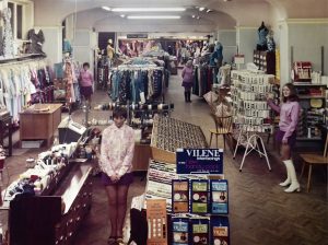 Vintage clothing shop interior with racks of colourful clothes, beauty products, and sewing supplies. Several women in 1960s fashion stand throughout the shop. The floor is herringbone wood, and the lighting is warm.