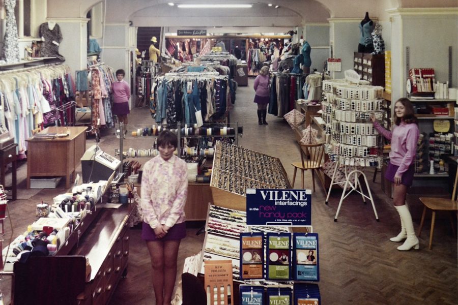 Vintage clothing shop interior with racks of colourful clothes, beauty products, and sewing supplies. Several women in 1960s fashion stand throughout the shop. The floor is herringbone wood, and the lighting is warm.