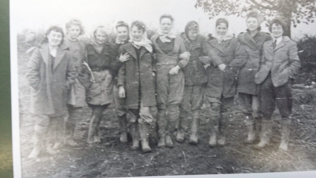 A vintage black-and-white photo shows ten people standing outdoors in two rows, dressed in coats and boots. The background has trees and uneven ground. The group is looking at the camera and smiling.