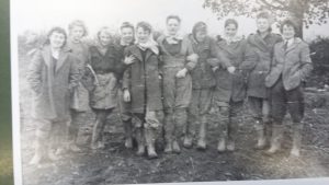 A vintage black-and-white photo shows ten people standing outdoors in two rows, dressed in coats and boots. The background has trees and uneven ground. The group is looking at the camera and smiling.