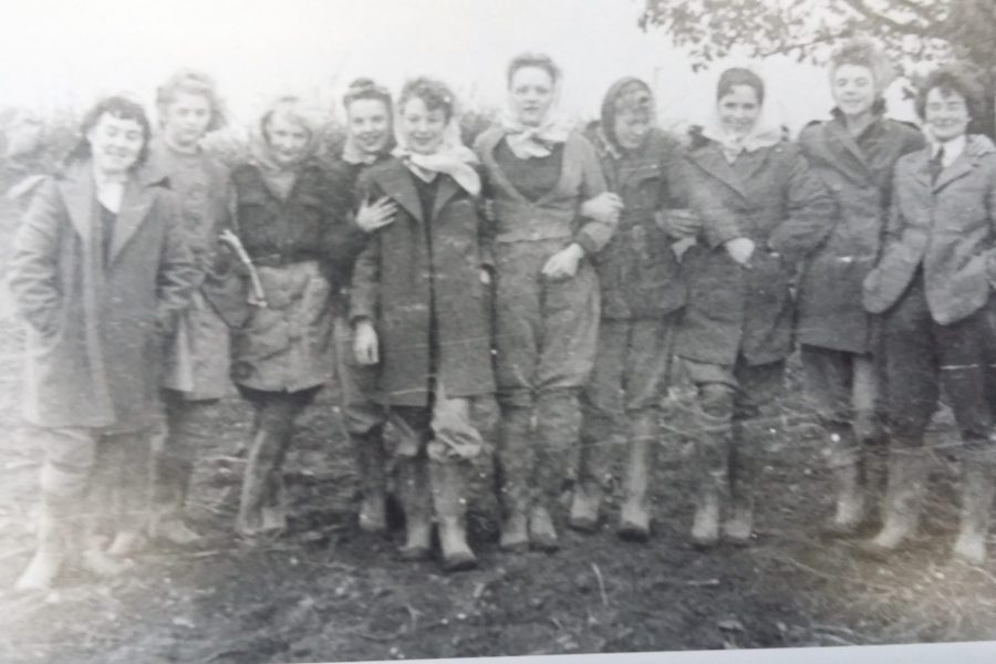 A vintage black-and-white photo shows ten people standing outdoors in two rows, dressed in coats and boots. The background has trees and uneven ground. The group is looking at the camera and smiling.