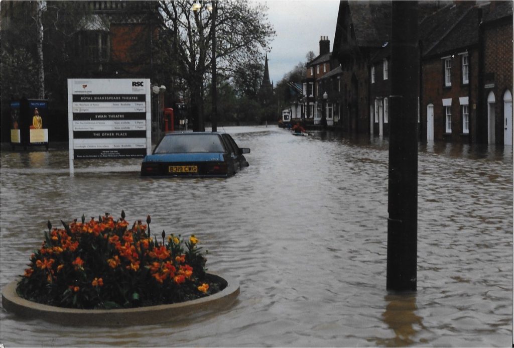 A flooded street with partially submerged cars, a flower bed in the foreground, and buildings along the roadside. A white sign for the Royal Shakespeare Theatre stands on the left.