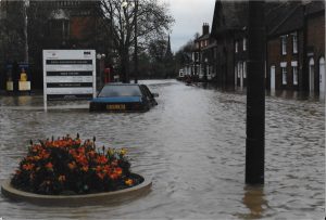 A flooded street with partially submerged cars, a flower bed in the foreground, and buildings along the roadside. A white sign for the Royal Shakespeare Theatre stands on the left.