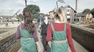 Four people in colourful costumes walk along a bridge; two women in green dungarees have gardening tools on their backs, ahead is a person with a white beard and another with a flamingo sculpture on their head. Buildings and sky in the background.