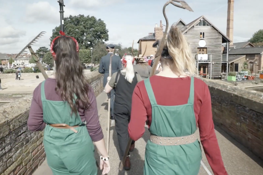 Four people in colourful costumes walk along a bridge; two women in green dungarees have gardening tools on their backs, ahead is a person with a white beard and another with a flamingo sculpture on their head. Buildings and sky in the background.